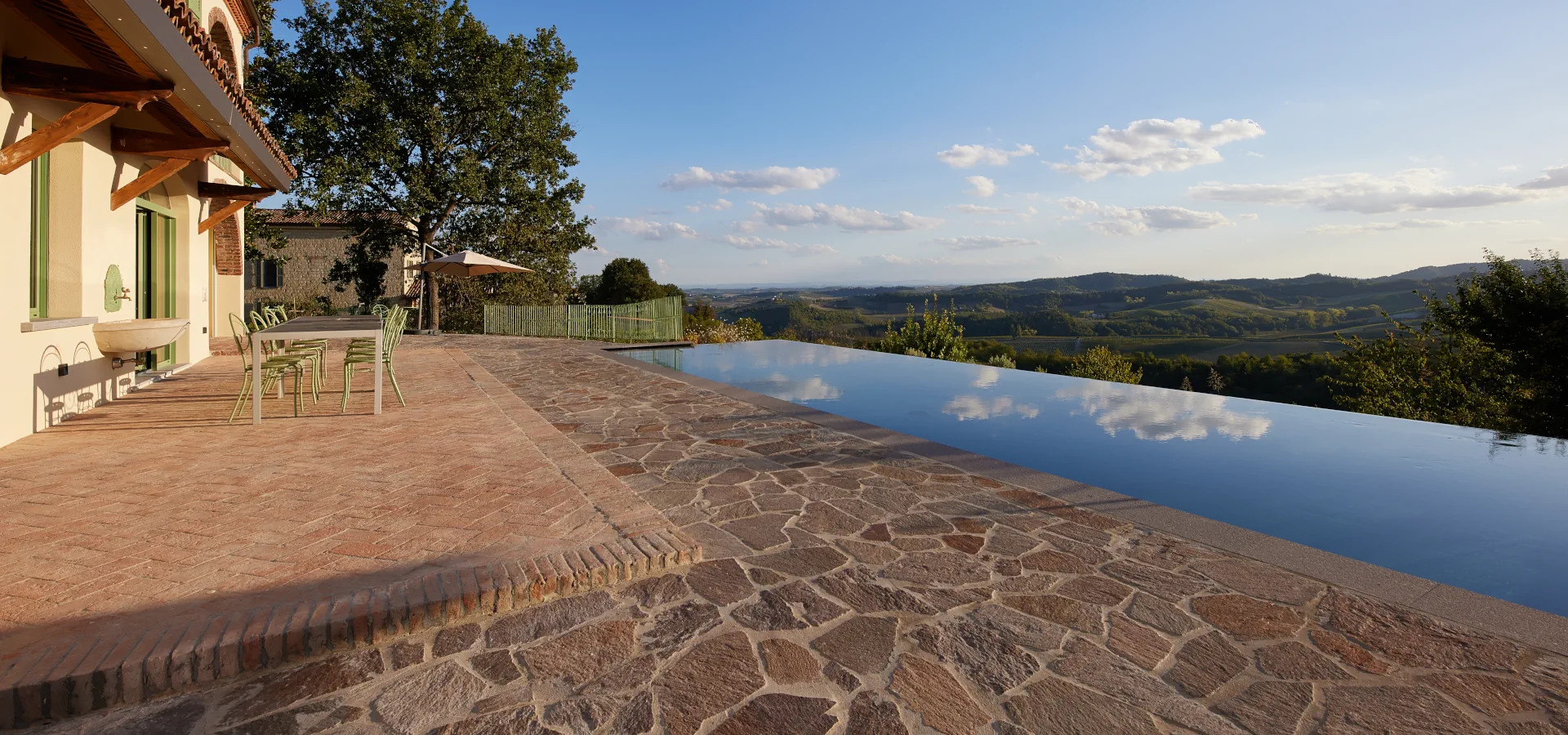 Villa Ozzano Monferrato - Vista della zona pranzo esterna con pavimento in cotto naturale, tettoia in coppi di recupero e piscina panoramica infinity con sfondo delle colline del Monferrato - Design FSA Filippo Selis Architetto
